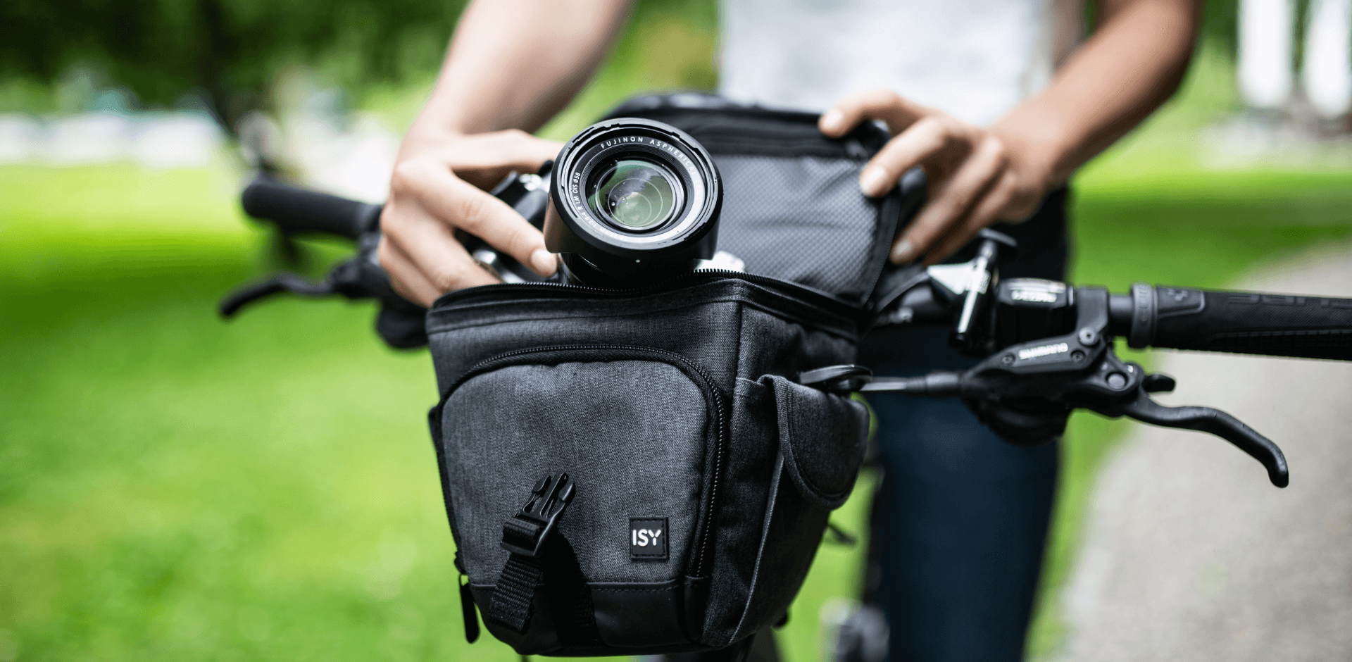 Jonge vrouw pakt een camera uit een cameratas van het merk ISY, tas zit vast aan haar fiets, in een park, close-upbeeld