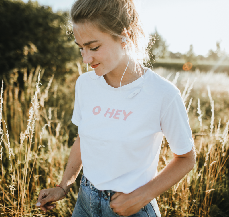 Une jeune femme écoute de la musique avec des écouteurs In Ear, dans un champ avec des hauts brins d’herbe, ambiance coucher du soleil d’été