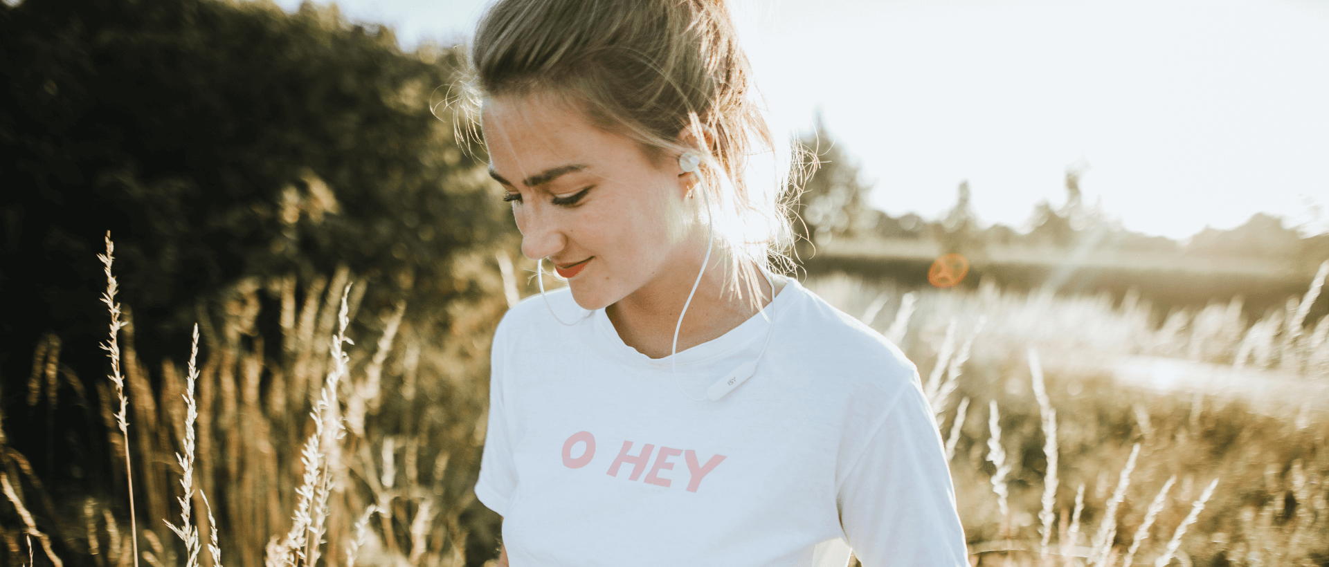 Jonge vrouw luistert naar muziek met in-ear oordopjes van het merk ISY, in een veld met hoge grashalmen, zomerse stemming, zonsondergang