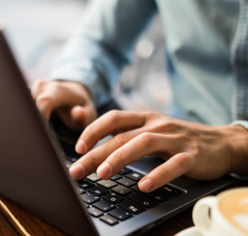 Junger Mann tippt auf einem Laptop von PEAQ, daneben eine volle Kaffeetasse, sitzt in einem Café, Nahansicht