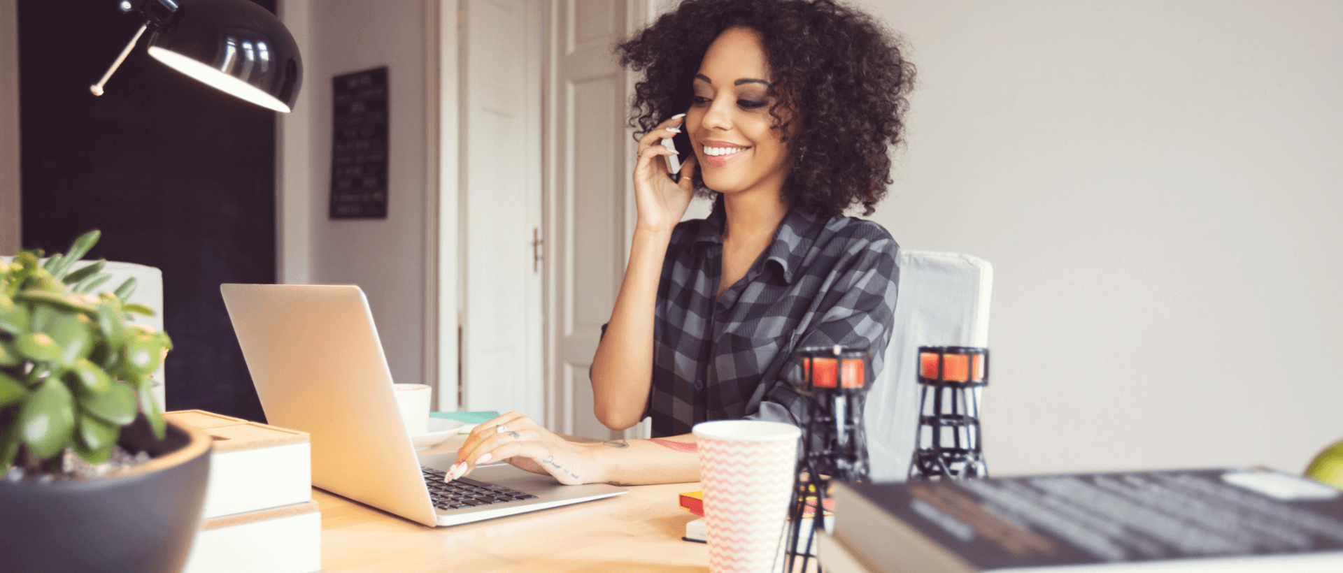 Una giovane telefona con il suo smartphone dalla sua postazione di lavoro, con l’altra mano scrive sul portatile, in ufficio